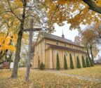 Widoczna fasada oraz nawa boczna kościoła jesienią w otoczeniu drzew z żółtymi liśćmi. Na pierwszym planie znajduje się stary, drewniany krzyż. Teren wokół kościoła pokryty opadłymi liśćmi.