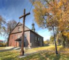 Kościół widziny z zewnątrz. Przed kościołem stoi drewniany krzyż. Wokół rosną drzewa. Jest jesień, liście są koloru żółtego.