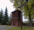 Dzwonnica stojąca obok kościoła. Przed dzwonnicą drewniany krzyż. 