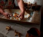 Gospodyni wyrabia ciasto na fafernuchy, mieszając składniki w drewnianym naczyniu. Na stole stoją figurki zwierząt z ciasta, tzw. byśki.