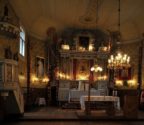 Wnętrze kościoła z widokiem na ołtarz główny. Z lewej strony znajduje się drewniana ambona. Ściany zdobią polichromie. We wnętrzu panuje półmrok rozświetlony lampkami.