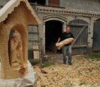 Józef Bacławski idzie przez podwórze, niosąc w rękach spory klocek drewna na rzeźbę. Na pierwszym planie kapliczka z Chrystusem Frasobliwym.