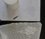 Wzór serwetki rozrysowany na papierze, szpulka białych nici, szydełko oraz koronka szydełkowa.