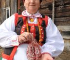 Halina Pajka przed drewnianym budynkiem gospodarczym. Jest ubrana w strój kurpiowski. W rękach trzyma dużą leluję wyciętą z papieru w kolorze bordowym.