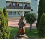 Budynek Centrum Kultury Kurpiowskiej w Kadzidle. Przed budynkiem stoi drewniana figura przedstawiająca świętego Rocha z psem.