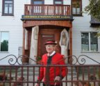 Henryk Kulesza w stroju kurpiowskim przed wejściem do ośrodka. Po obu stronach drzwi stoją kapliczki wyrzeźbione w pniach drzew.