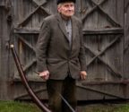 Jan Maliszewski stoi na podwórku przed drewnianym budynkiem. W ręce trzyma ligawkę zawieszoną na pasku.