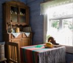 Fragment izby. Ściany są pomalowane na niebiesko. W rogu stoi kredens z przedmiotami kuchennymi. Przed kredensem, przy oknie, stoi stół i krzesło. Obok drewniane sprzęty używane w gospodarstwie domowym. 