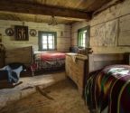 Wnętrze jednej z chat, W izbie stoją dwa łóżka, skrzynia wianna, kołyska dla dziecka i krosna. Na ścianie święty obraz i haftowana makatka.