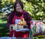 W ogrodzie stoi Anna Szpura, trzymając w rękach korowaj ozdobiony parą łabędzi z ciasta. W tle zielrń drzew, kwiaty, ul.