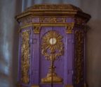 Drewniane tabernakulum w kolorze fioletowym ze złoconymi zdobieniami. Na froncie przedstawienie monstrancji.