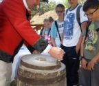 Mężczyzna w stroju kurpiowskim pokazuje grupie dzieci jak mleć ziarno na mąkę za pomocą żarna, czyli obracanego siłą rąk kamienia. Dzieci przyglądają się z zainteresowaniem.