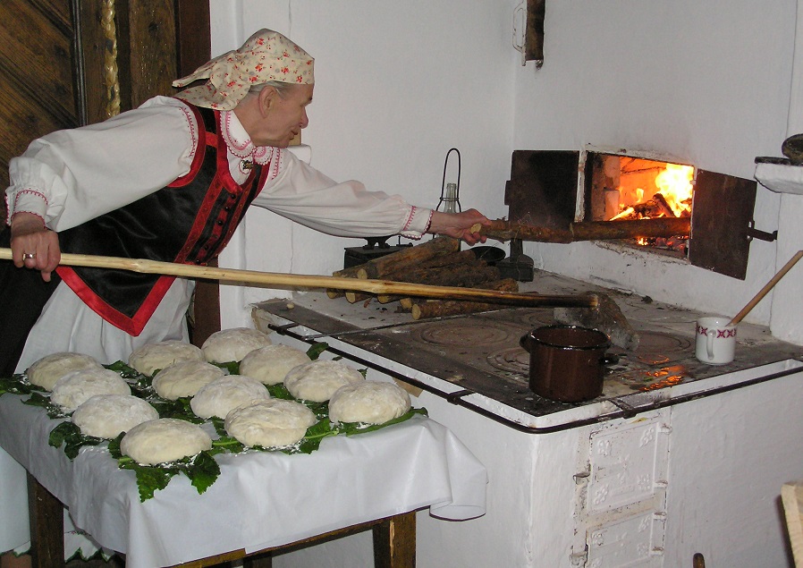 Kobieta dorzuca drewno do ognia w tradycyjnym piecu do wypieku chleba. Na stole leżą surowe bochenki przygotowane do pieczenia.