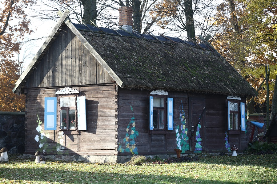Chata kurpiowska z krytym strzechą dachem. Okiennice są pomalowane na niebiesko. Na ścianach wymalowane są malwy.