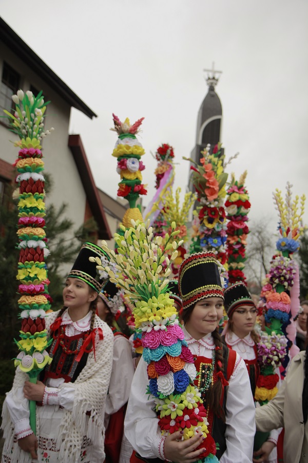 Grupa dziewcząt z strojach ludowych idzie w procesji podczas Niedzieli Palmowej. Dziewczęta niosą kolorowe kurpiowskie palmy.
